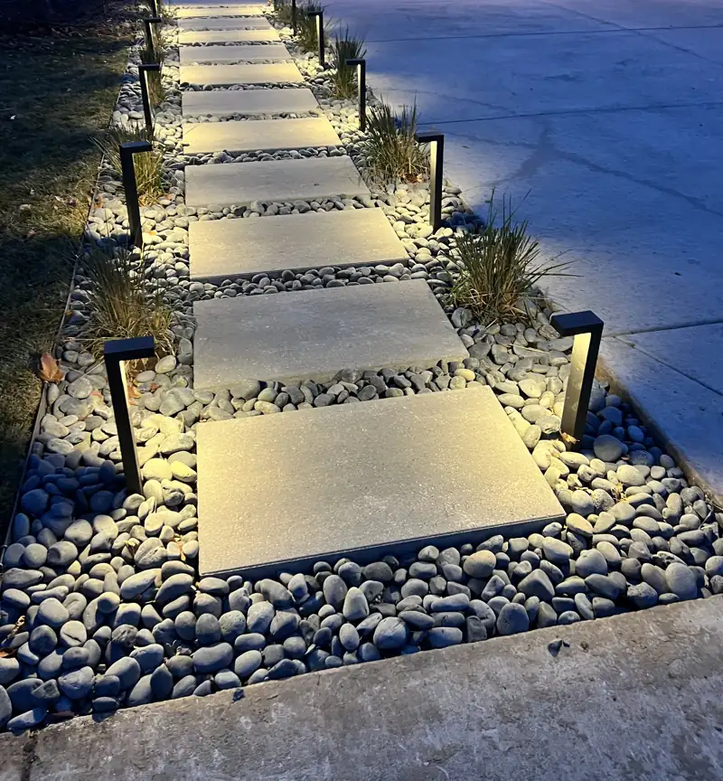 Black bollard landscape path lights illuminating a large-format paver stepping-stone walkway with river rock and ornamental grasses at dusk