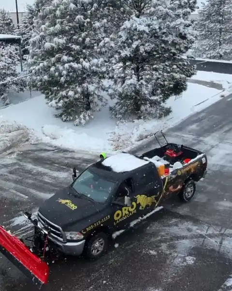 Oro Landscape snow removal service plow truck clearing a snowy lot, with a snow blower loaded in the truck bed